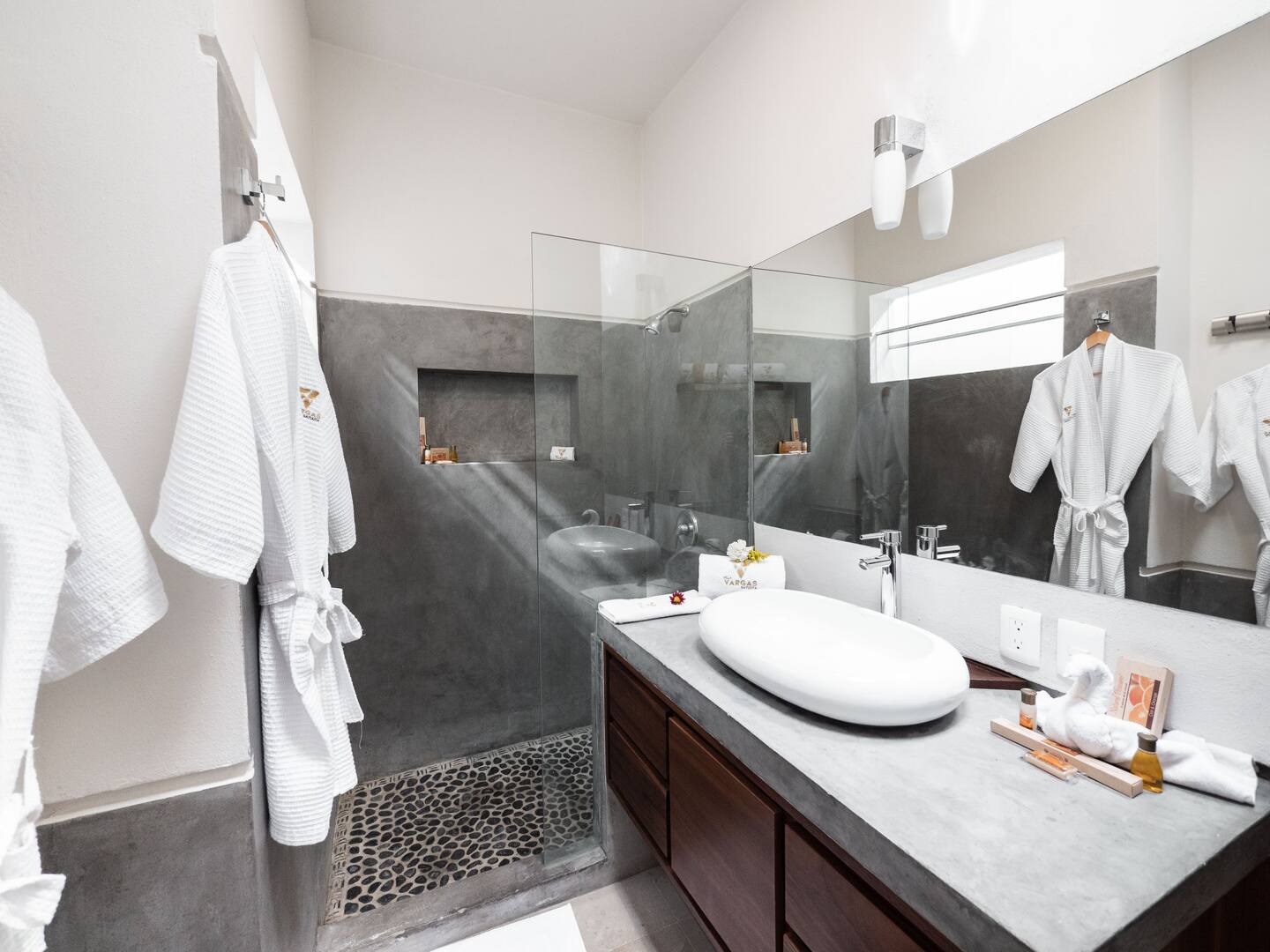 The ensuite bathroom off the master bedroom at Villa Vargas Sayulita, with white robes on the wall, a long polished concrete vanity with a vessel sink and a frameless glass shower with a black pebbled floor