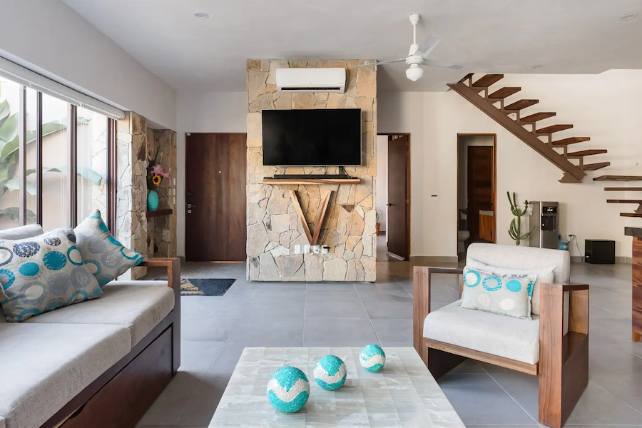 The main living room at Villa Vargas Sayulita with sofas, a stone feature wall carrying a flat-screen TV, and the floating staircase behind