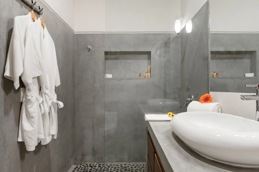 The ensuite bathroom off the twin bedroom at Villa Vargas Sayulita, with robes hanging beside a walk-in polished concrete shower with a pebbled floor and a concrete vanity with a vessel sink