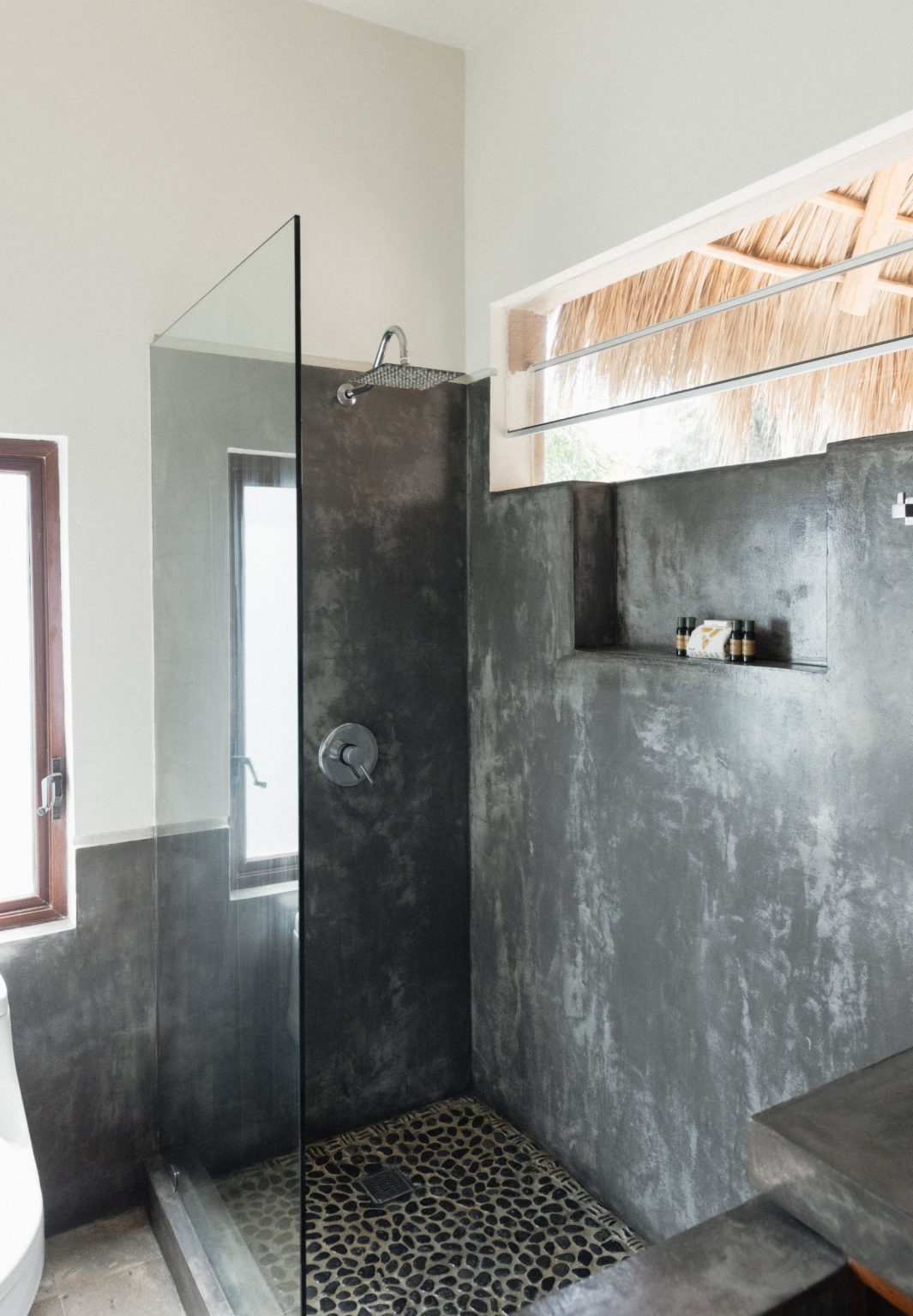 The ensuite bathroom off the two-queen bedroom at Villa Vargas Sayulita: a glass-panelled walk-in rain shower in polished concrete with a black pebbled floor, a recessed shelf of toiletries and a clerestory window onto the palapa roof