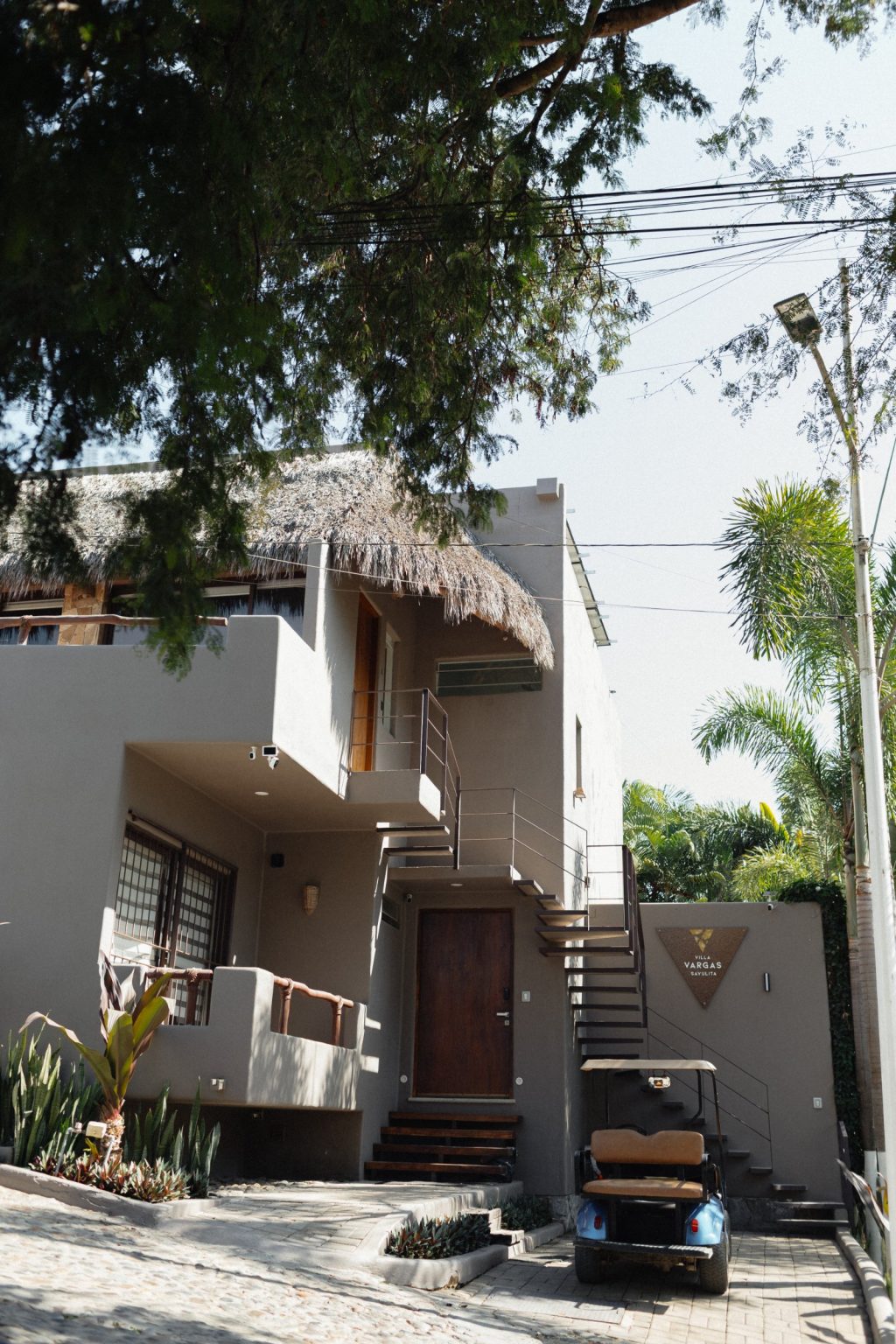 The front of Villa Vargas Sayulita from the cobbled street, with a hardwood front door, the exterior stair to the upper entrance, the palapa roof above and the villa sign on the wall