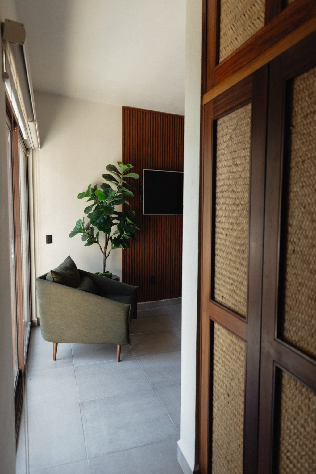 The entry into the master bedroom at Villa Vargas Sayulita, past woven-panel doors to a green sofa, a tall plant and a television on the slatted hardwood wall