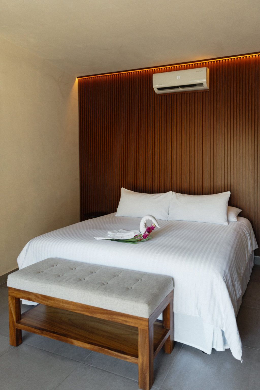 The master bedroom at Villa Vargas Sayulita: a king bed made up in white linen against a full-height slatted hardwood headboard wall lit from behind, with an upholstered bench at the foot of the bed