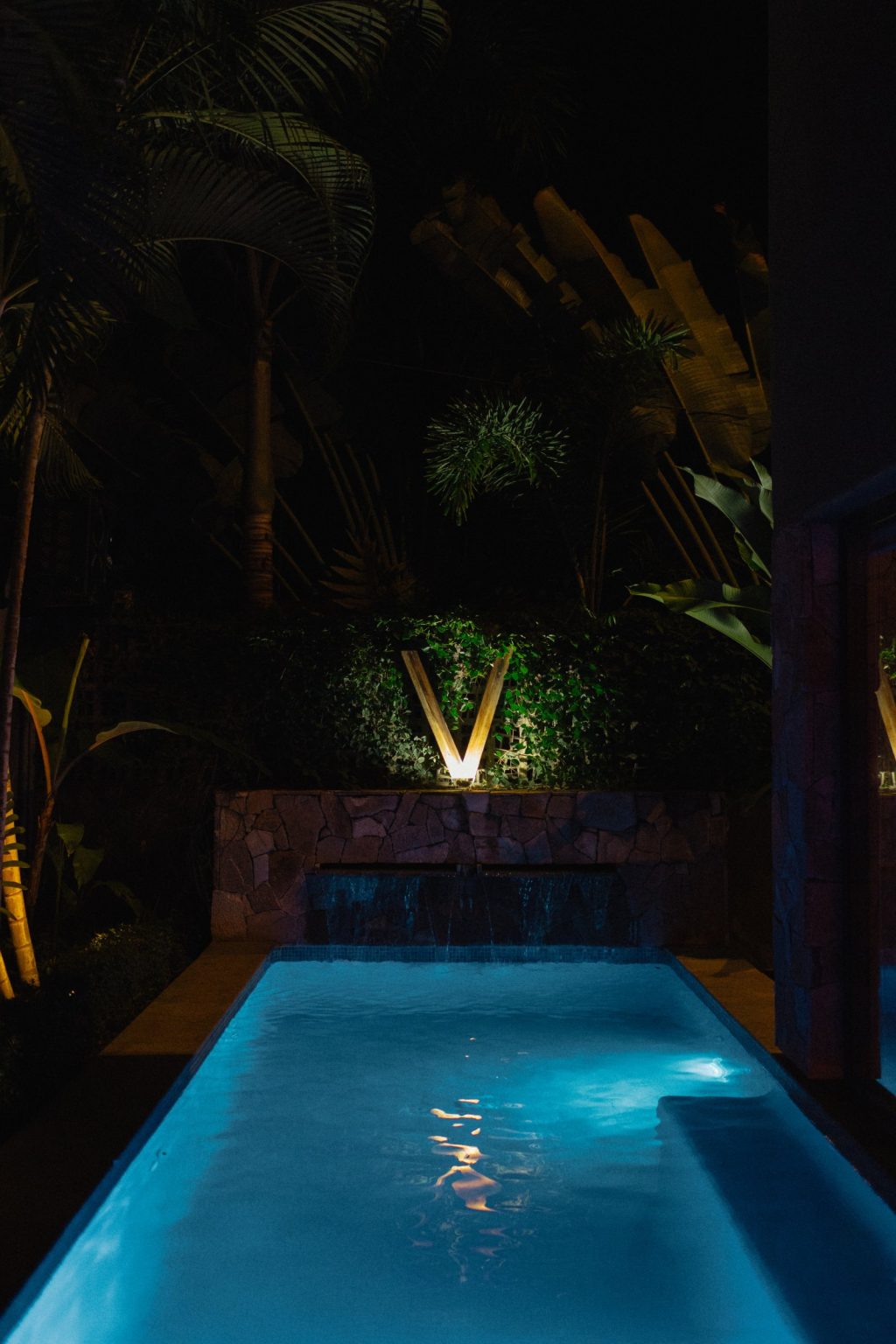 The private pool at Villa Vargas Sayulita lit blue at night, with the stone waterfall wall and the illuminated villa mark in the planting behind it