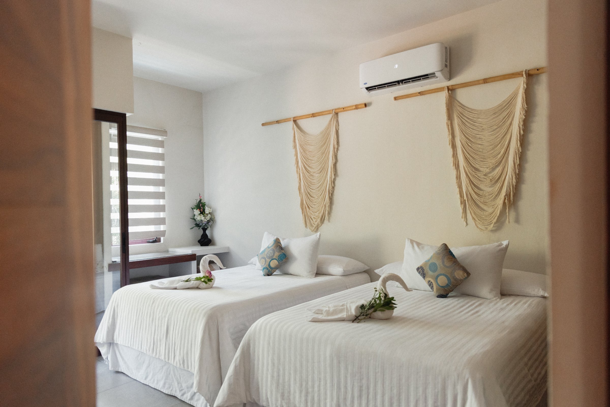 The upstairs bedroom at Villa Vargas Sayulita seen from the doorway: two queen beds made up in white linen under a pair of macrame wall hangings on a bamboo rod, with air conditioning above